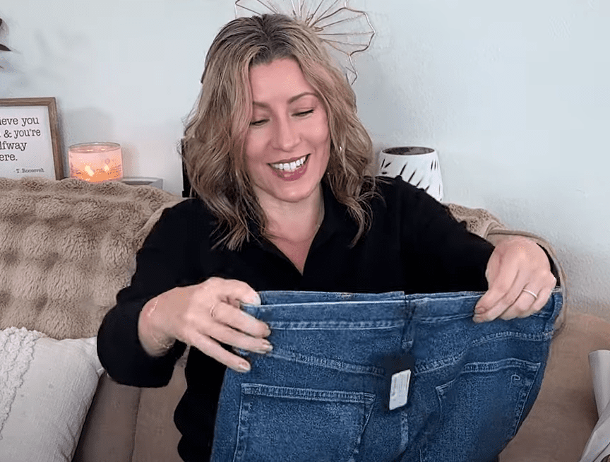 A person with wavy hair sits comfortably on a couch, smiling in a black top. Theyre holding up a pair of stylish sweatpant jeans with a tag. In the background, theres a lit candle and a decorative sign on a shelf.